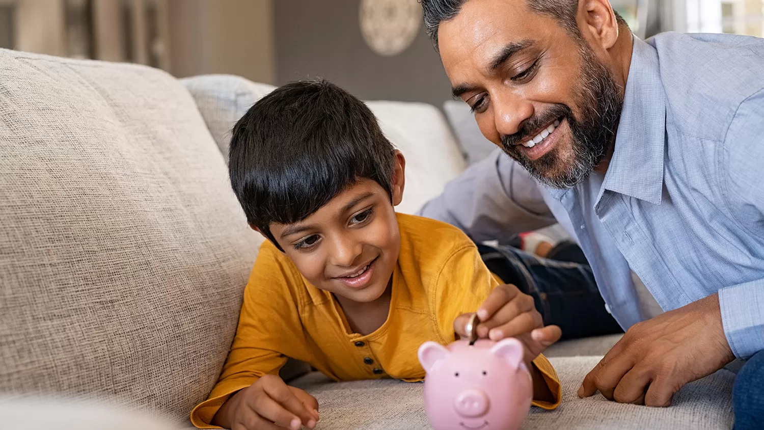Son and father laying on a couch while son drops a coin in a piggy bank
