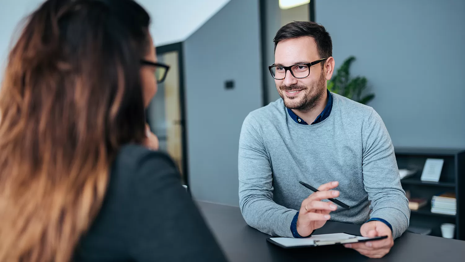 Male sitting at a table while holding a pen and clipboard as he is smiling and talking to a female