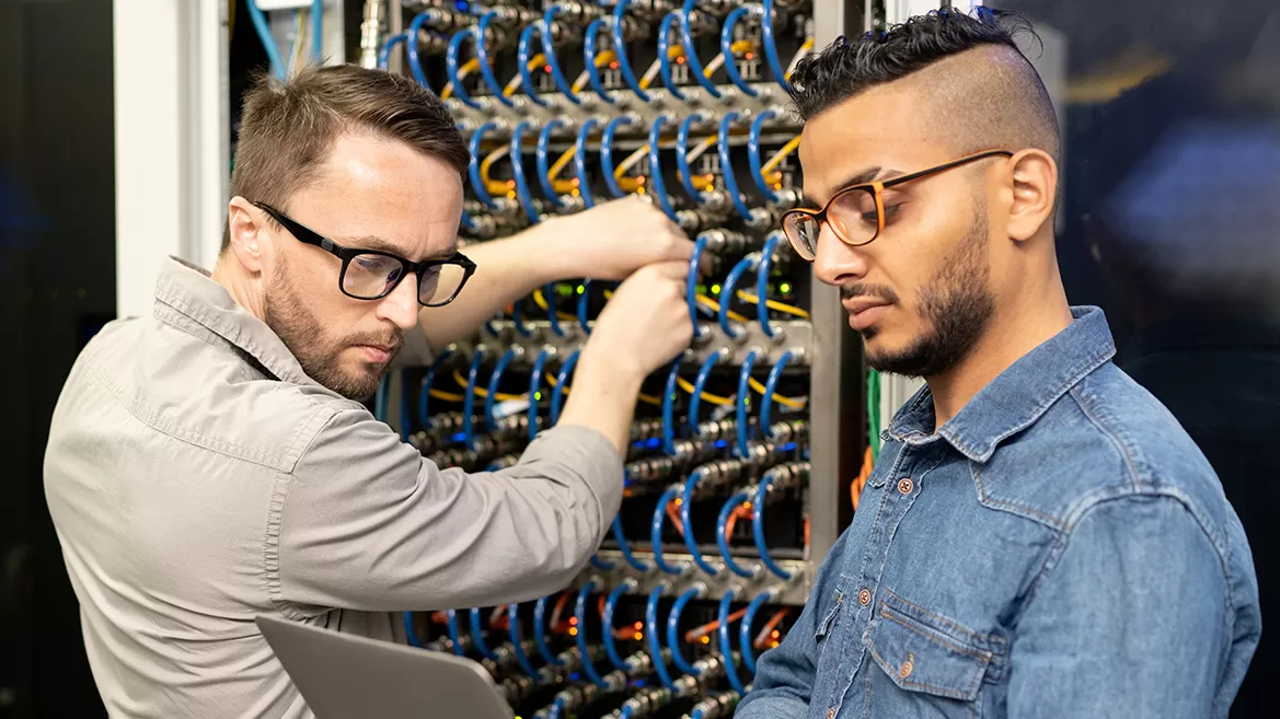 Two men working on a server cabinet while examining information on a laptop